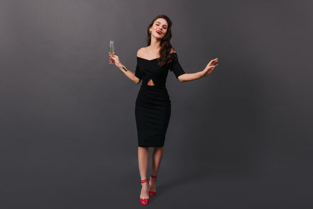 衣服身着黑色合身连衣裙和高跟鞋的女子手拿香槟酒杯在黑色背景上摆姿势的完整照片节日年轻时尚