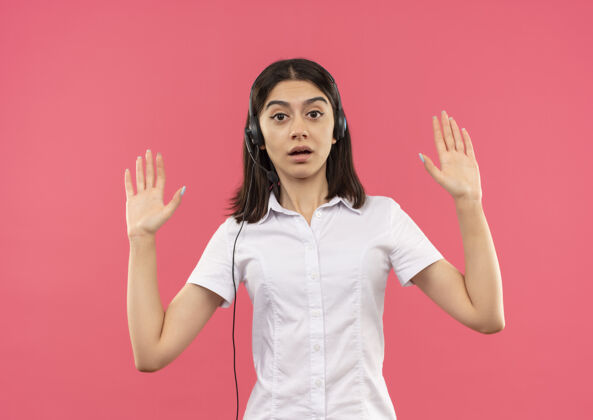 人穿着白衬衫 戴着耳机的年轻女孩站在粉色的墙上 举手投足 看上去很惊讶女性提高耳机