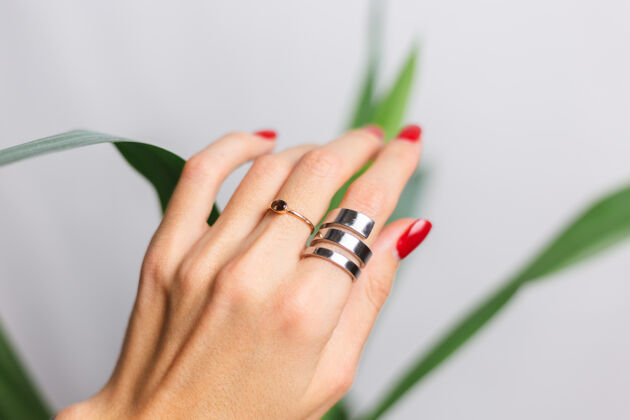 新鲜女人的手上有红色的指甲和两个指环 放在美丽的绿色棕榈叶上后面是灰色的墙手成人自然