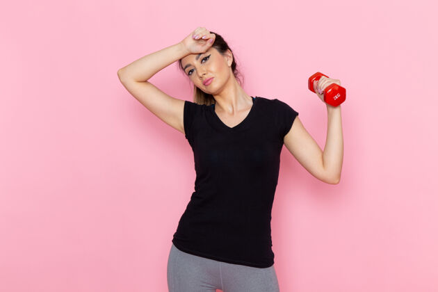 运动正面图：年轻女性手持哑铃在浅粉色墙上运动员运动锻炼成人轻漂亮