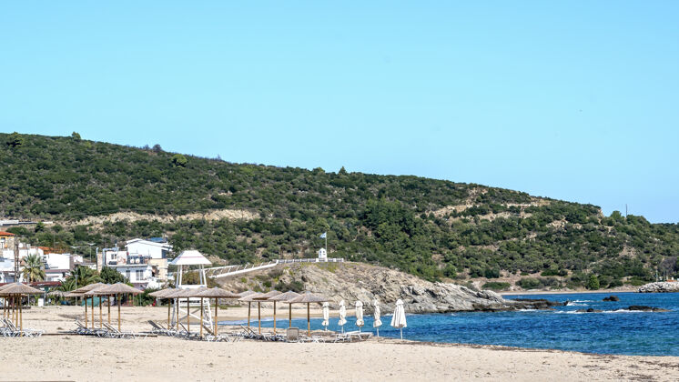 岩石爱琴海海岸左侧有建筑物 岩石 带日光浴床的雨伞 灌木丛和树木 蓝色的海水和希腊萨蒂的小山海海岸悬崖