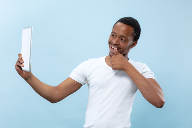 长相半身特写 蓝色背景 白色衬衫的非洲裔美国年轻男子人类情感 面部表情 广告 销售 概念使用平板电脑进行自拍 视频日志 交谈成人肖像选择