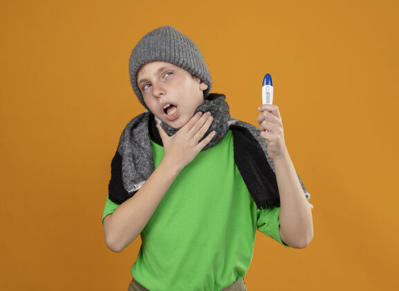 小生病的小男孩穿着绿色t恤 戴着暖和的围巾 戴着帽子 体温表显示他身体不适 不舒服 手放在喉咙上 站在橙色的墙上手体温计橙色