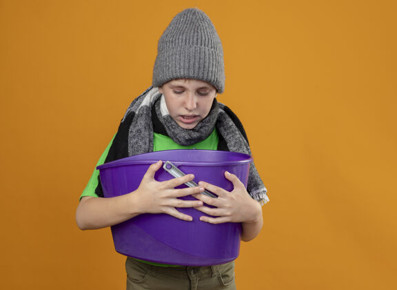 帽子生病的小男孩穿着绿色t恤 戴着暖和的围巾 戴着帽子 手里拿着垃圾 站在橙色的墙上感到恶心小男孩围巾