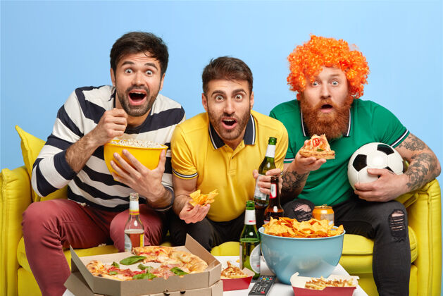 沙发兴奋的三个男朋友聚精会神地看电视屏幕 兴致勃勃地看足球比赛 在宽敞的客厅沙发上摆姿势 吃爆米花球迷胡须食物