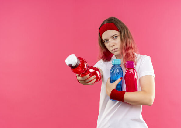 水戴着头巾的年轻运动型女人拿着几瓶水 站在粉红色的墙上献上一瓶水头带女人运动