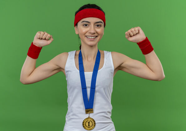 脖子戴着头带 脖子上戴着金牌的年轻健身女士举起拳头 像一个站在绿色墙壁上自信微笑的胜利者喜欢周围奖牌