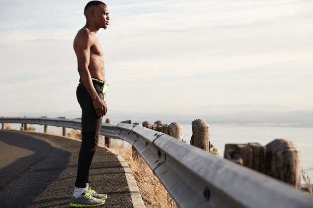 运动员皮肤黝黑的运动型男子侧身拍摄 在城外接受健身训练 穿着运动服 认真自信地眺望远方 站在柏油路上 感觉健康人与运动运动鞋景观呼吸