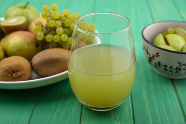 果汁前视图：一杯软饮料 绿色背景 盘子上有绿色苹果 猕猴桃 绿色葡萄和梨猕猴桃饮料玻璃