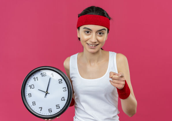 运动装身穿运动服的年轻健身女 头戴头巾 手持挂钟 指着摄像机 脸上带着微笑 站在粉色的墙上墙头带女人