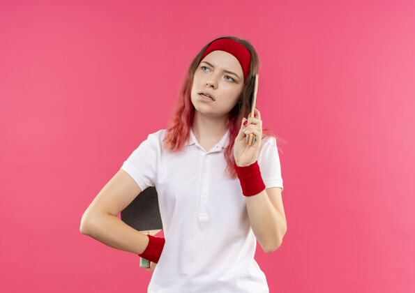 脸戴着头巾 拿着两个乒乓球球拍的年轻运动女郎站在粉红色的墙上 脸上带着沉思的表情望向一边年轻人站着球拍