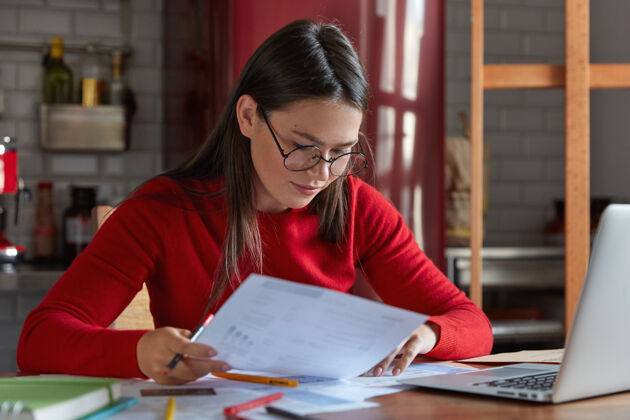 注意项目经理戴着眼镜和红色juumper的水平镜头 仔细看文件 思考如何吸引客户和增加收入 用便携式笔记本电脑对着厨房内部摆姿势自由职业者文件年轻人
