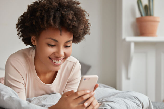 卧室快乐的黑人美国妇女躺在床上用现代手机 阅读愉快的评论下她的照片在社交网络通过智能手机 享受床上的时间和互联网在舒适的房间独自床非洲非洲