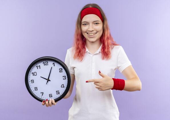 站着年轻的运动型女人拿着挂钟 手指着挂钟 站在紫色的墙上欢快地笑着运动运动女人