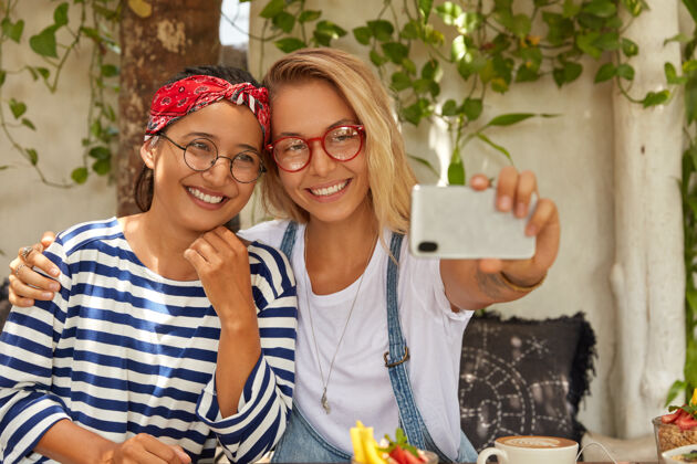 眼镜友好的多民族女性在手机中拥抱和摆姿势 自拍肖像 在咖啡馆消遣 吃甜点 戴圆眼镜 享受娱乐时间 满意的东西相聚积极伴侣