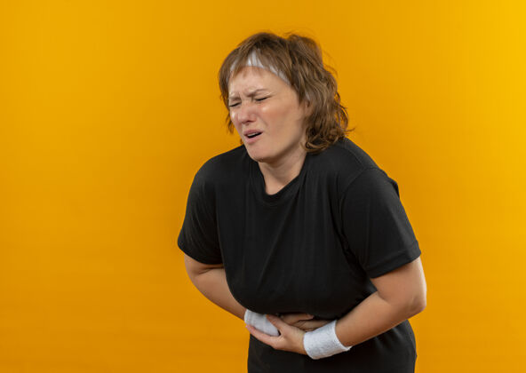 痛苦中年运动女性 身穿黑色t恤 头箍看起来不舒服 站在橘色的墙上摸着肚子 疼痛难忍中间不适姿势