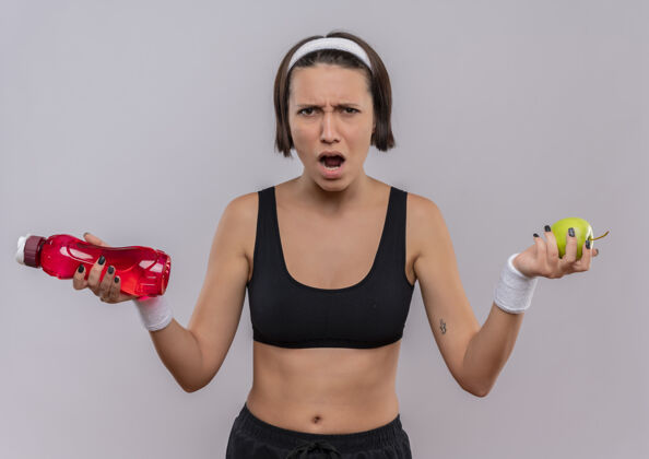 运动穿着运动服的年轻女健身者拿着一瓶水和绿苹果站在白墙上怒气冲冲地喊着运动员人看着