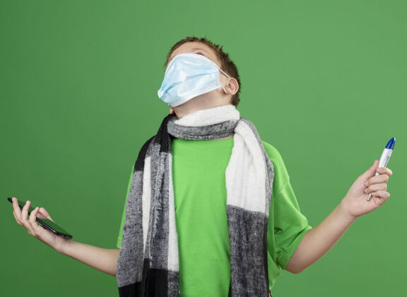 抱着生病的小男孩穿着绿色t恤 脖子上围着暖和的围巾 戴着护面面具 拿着计价器和智能手机 站在绿色背景下显得很沮丧男孩周围围巾