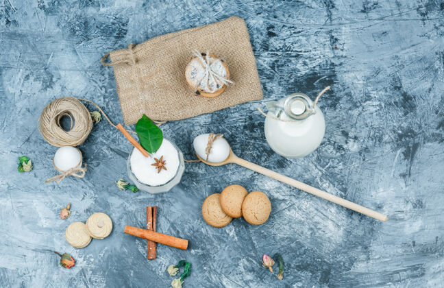 冷俯视图：一罐牛奶和一杯酸奶 还有勺子 饼干 鸡蛋 线绳 肉桂和一棵植物 放在深蓝色大理石表面水平饮食早餐鸡蛋