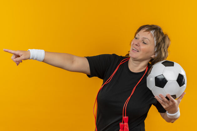 抱着中年运动女性 身穿黑色t恤 头带 手持足球 手指指向一边 微笑着站在橙色的墙上头带人运动