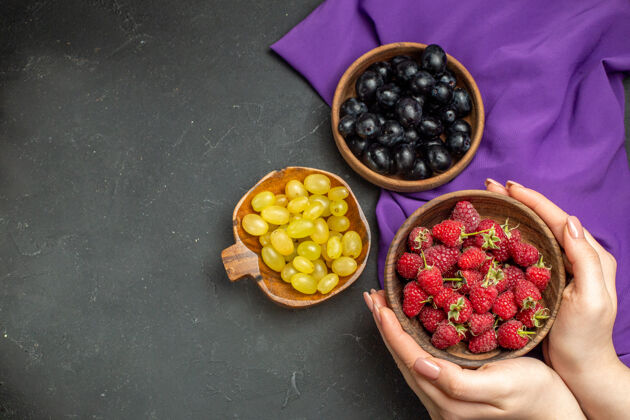 顶部顶视图树莓在女性手中碗黑色和黄色葡萄在碗紫色披肩黑暗孤立的表面自由空间碗浆果覆盆子