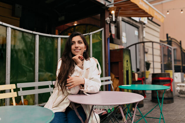 青少年浪漫害羞的长发女孩穿着白大褂坐在户外的法式咖啡馆里等待约会自信表情微笑