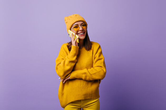 紫色满意的女人 留着一头长发 一边用智能手机和好友聊天 一边开心地笑着一个戴着时髦帽子 穿着黄色衣服 面带微笑的学生想在孤立的紫色上做一个快照肖像欢呼毛衣肖像