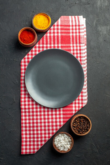黑色圆盘子顶视图黑色圆形盘子上的红色和白色方格餐巾碗与姜黄红胡椒粉海盐黑胡椒在黑暗的桌子上姜黄厨房用具炒锅
