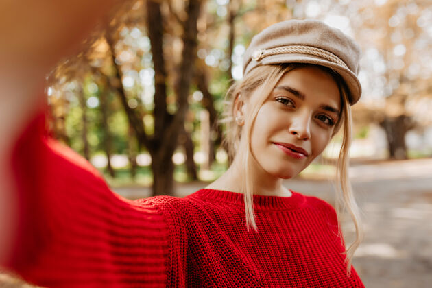 成人一位穿着红色毛衣的迷人金发女郎在秋季公园自拍的特写照片自然妆容的美丽年轻女子戴着时髦的轻便帽子在户外摆姿势微笑户外毛衣