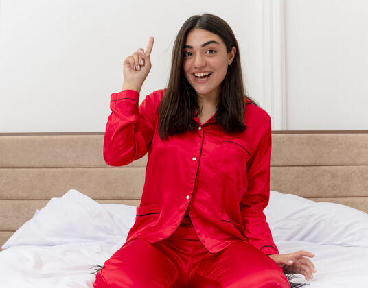 新穿着红色睡衣的年轻美女坐在床上看着相机开心地笑着在灯光背景下展示食指在卧室内部有新的想法漂亮睡衣看