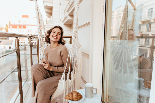 快乐快乐的白人女孩拿着美味的羊角面包坐在露台上一张年轻女子在阳台上吃早餐的照片休闲户外享受