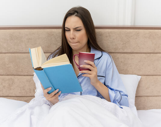 躺着穿着蓝色睡衣的年轻漂亮女人躺在床上 手里拿着书和咖啡 在灯光的背景下 带着怀疑的表情看着卧室内部卧室表情漂亮