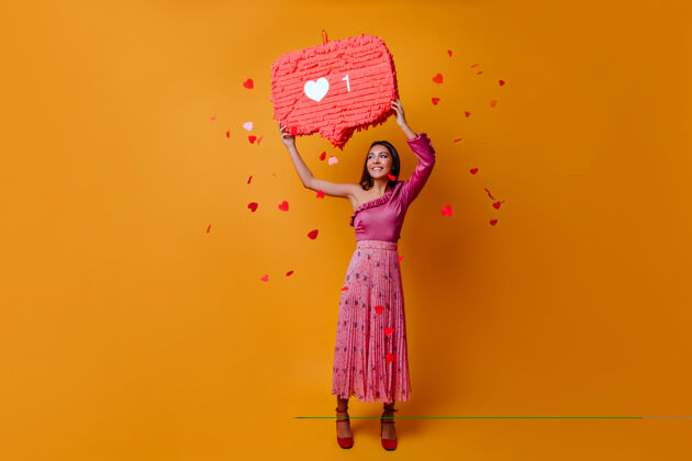 连接23岁的幸运迷人的女人拿着instagram的like标志 在橙色的墙上摆满了五彩纸屑喜欢信息舞蹈