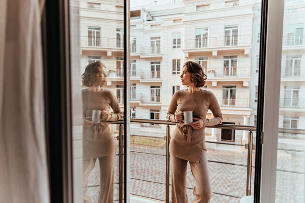 城市穿着考究的卷发女人站在大窗户边喝着咖啡美丽的白种女人喝茶看街的照片摩天大楼女人室内