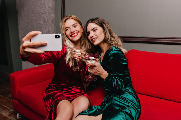 积极黑发女人一边喝酒 一边和朋友自拍放松的女孩坐在红色沙发上的室内写真室内积极黑发