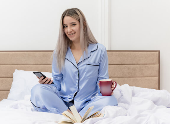 微笑穿着蓝色睡衣的年轻美女坐在床上 用智能手机喝着咖啡 在卧室的室内灯光背景下微笑着蓝色卧室坐着