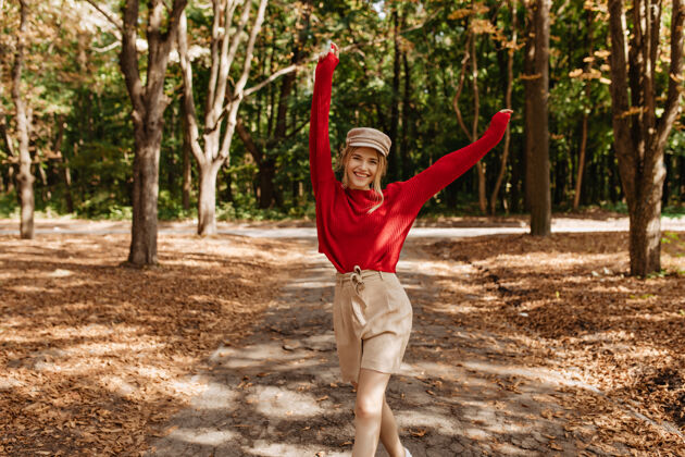 帽子穿着漂亮的红色毛衣和米色短裤的快乐金发女人在秋天的公园里跳舞时髦的年轻女人在户外的好天气里快乐地摆姿势树叶花朵裙子