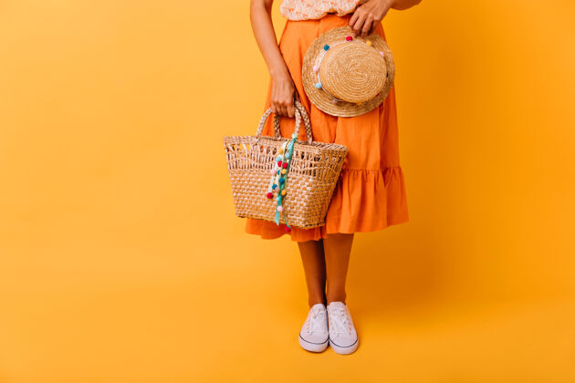 欢乐穿着橘色裙子和白色鞋子的晒黑女孩站在黄色的舞台上漂亮的女模特戴着时髦的帽子在摄影棚摆姿势欢呼裙子帽子