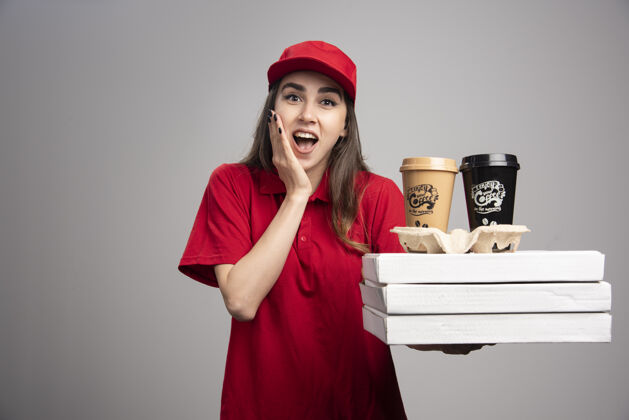 制服快乐的送货员在灰色的墙上拿着比萨饼和咖啡杯承运人咖啡人