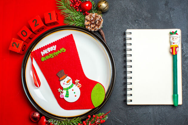 数字新年背景俯视图 圣诞袜子放在餐盘上 装饰配件杉木树枝和数字放在红色餐巾上 笔记本旁边是黑色桌子上的钢笔餐盘笔记本餐巾