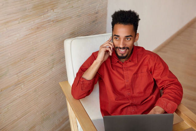 男士快乐的年轻黑发胡子黑皮肤的男性微笑着愉快的电话交谈 坐在椅子上与笔记本电脑在米色内饰情绪表情电话