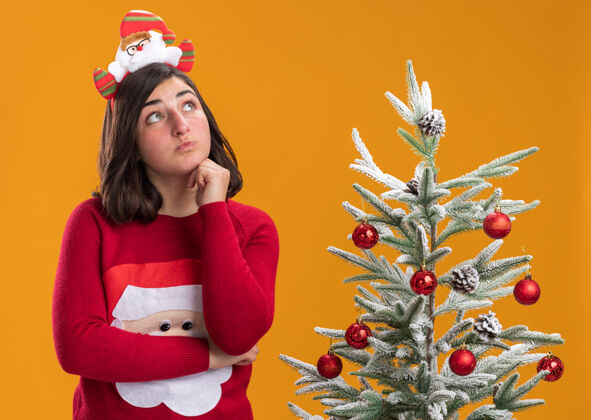 穿着穿着圣诞毛衣戴着滑稽头带的年轻女孩站在橙色墙上的圣诞树旁 困惑地抬头望去年轻头带困惑