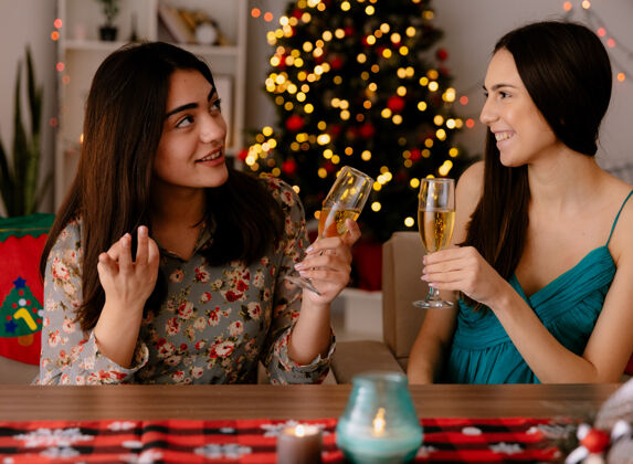 享受高兴的年轻姑娘们端着香槟酒杯坐在餐桌旁 在家里享受圣诞节时光坐着圣诞快乐圣诞节