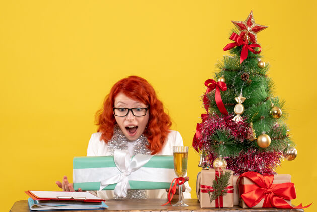 前面前视图：女医生拿着圣诞礼物和黄色背景上的圣诞树坐着圣诞节圣诞树漂亮