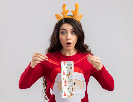 新年好奇的年轻漂亮女孩戴着驯鹿鹿角头带和圣诞老人毛衣拿着圣诞礼物包打开看圣诞老人礼物戴