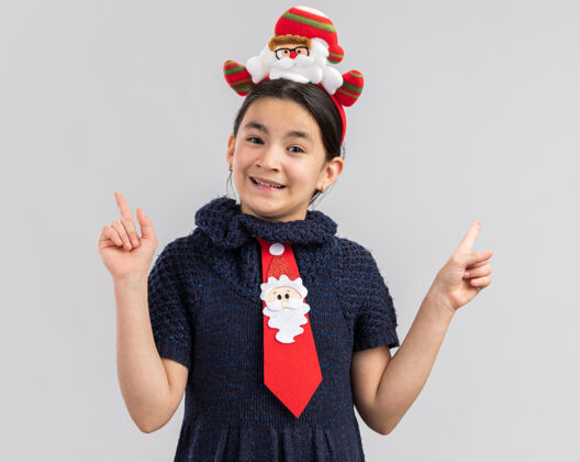 圣诞快乐小女孩穿着针织连衣裙 戴着红色领带 头上戴着有趣的圣诞戒指 快乐而积极地微笑着 露出食指红色展示新年