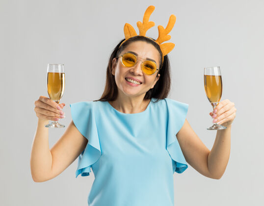快乐身着蓝色上衣的快乐年轻女子 戴着滑稽的鹿角边 戴着黄色眼镜 手里拿着两杯香槟 欢快地笑着戴着圣诞快乐两个