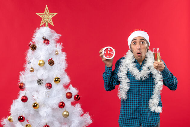 站着一个戴着圣诞老人帽子 拿着一杯葡萄酒和一个闹钟站在圣诞树旁的年轻人震惊了圣诞老人钟圣诞老人