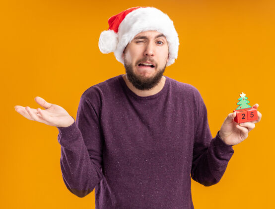 数字一个穿着紫色毛衣 戴着圣诞帽的年轻人站在橙色的墙上 展示着25号玩具立方体 困惑而不高兴地耸耸肩站着五毛衣
