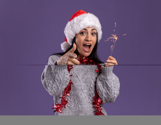 圣诞节令人印象深刻的年轻白人女孩戴着圣诞帽 脖子上戴着金箔花环 手持节日火花灯 隔离在紫色的墙上 留有复制空间花环指向闪闪发光